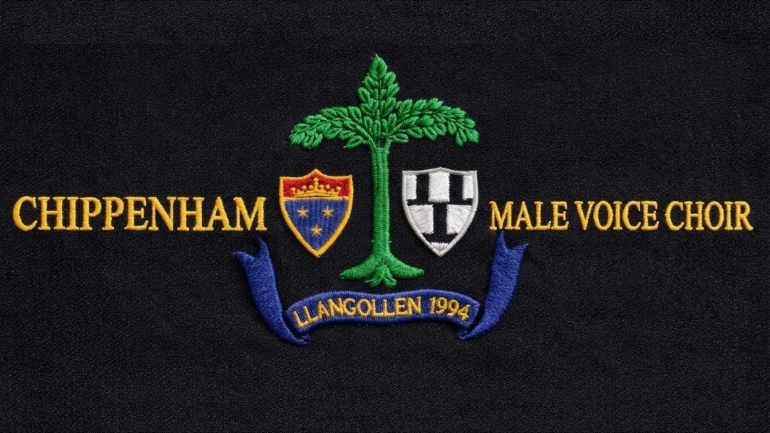 Embroidered heraldic logo identifies Chippenham Male Voice Choir. Design features green tree, blue-and-gold shield, silver-and-black shield, blue ribbon, and “LLANGOLLEN 1994” milestone banner.