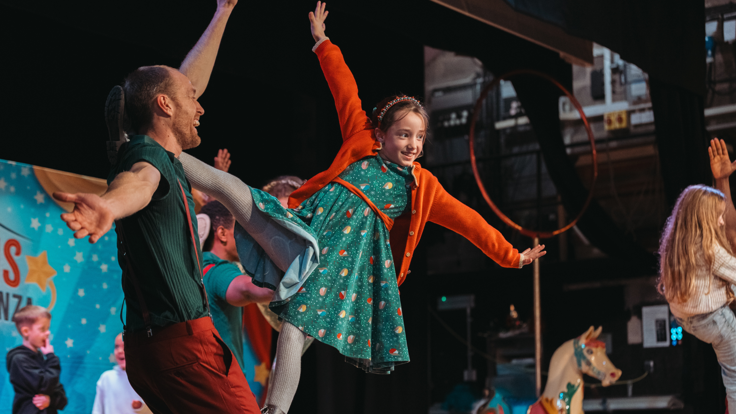 Photograph shows a young girl in a green patterned dress and orange cardigan performing a acrobatic move with an adult male partner on a colourful stage. Raised arms, extended legs, circus-themed backdrop, carousel horse, and audience members emphasize a lively theatrical performance.